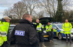 A group of men in high vis jackets are seen standing in a semi circle. There are police cars in the background.