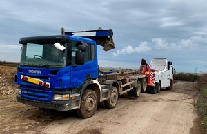 Image shows large tipper truck to left being lifted by its rear and towed away by heavy duty recovery truck