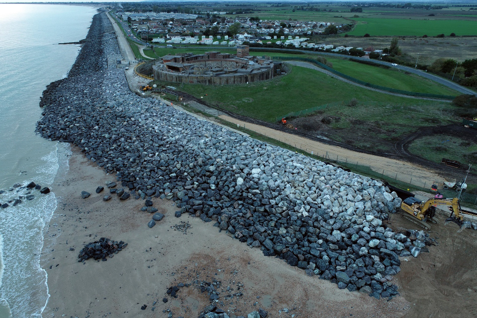 Image shows an angled bank of rocks on the shoreline protecting a large, circular concrete structure and buildings behind
