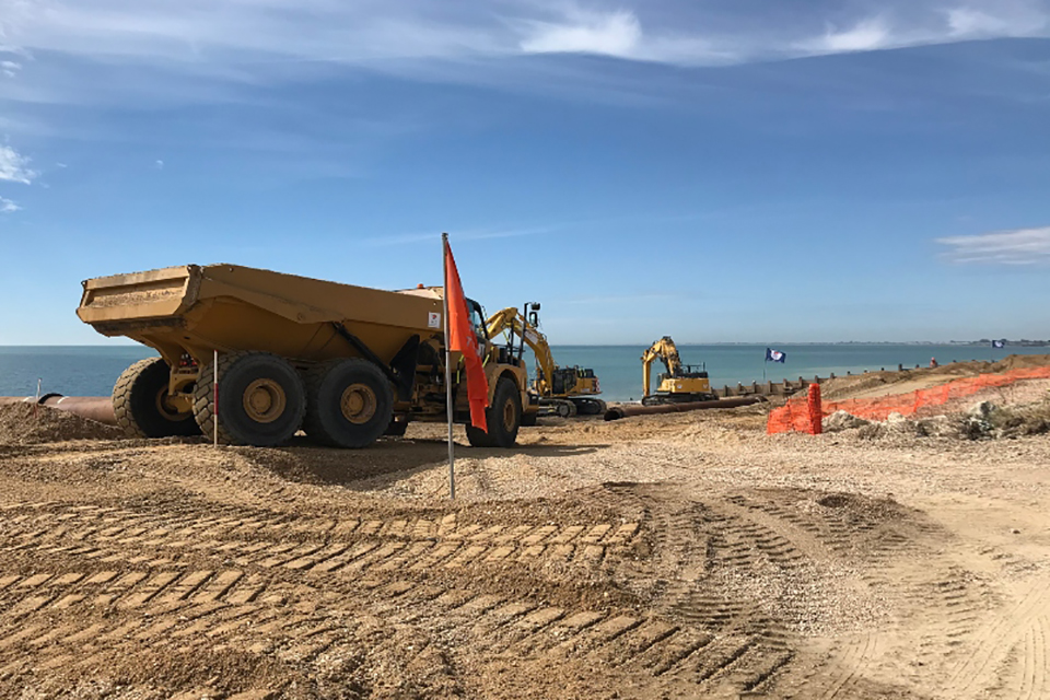 Image shows large, yellow dumper truck to the left travelling along a beach with two excavators ahead