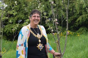 Lady with floral jacket and mayoral necklace holds a tree in hand ahead of planting in parkland in background.