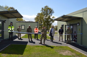 9 men stand between 2 new military buildings that are joined together by a ribbon. Lt Gen Richard Nugee cuts the centre of the ribbon.