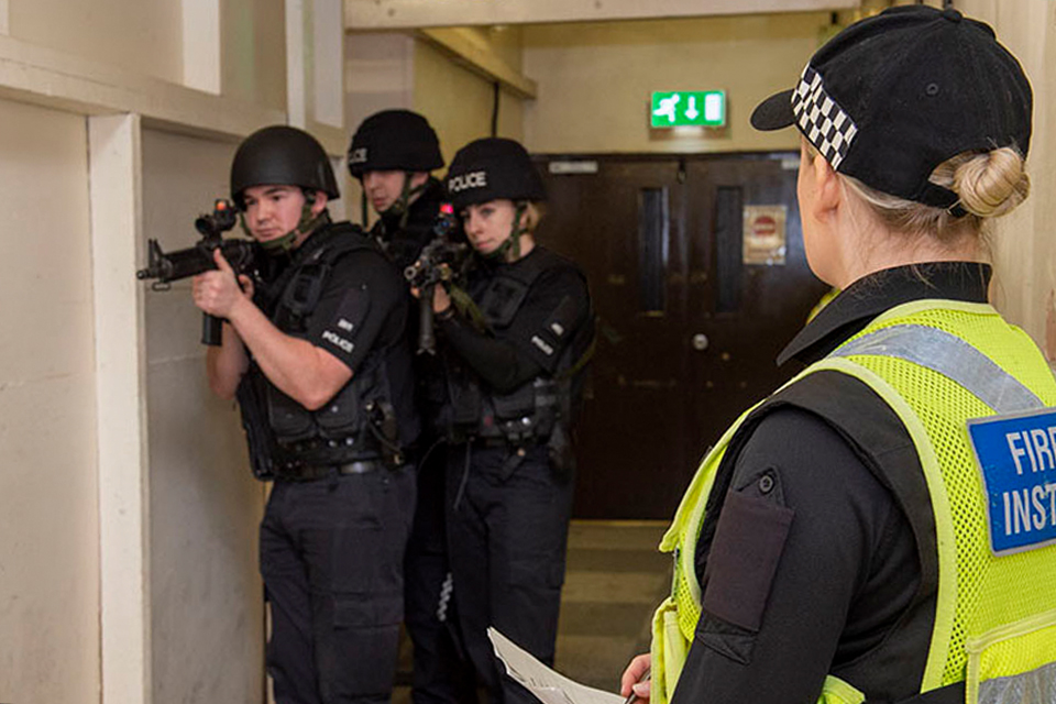 3 uniformed MDP police officers inside a building hallway holding firearms. A firearms training officer stands by.