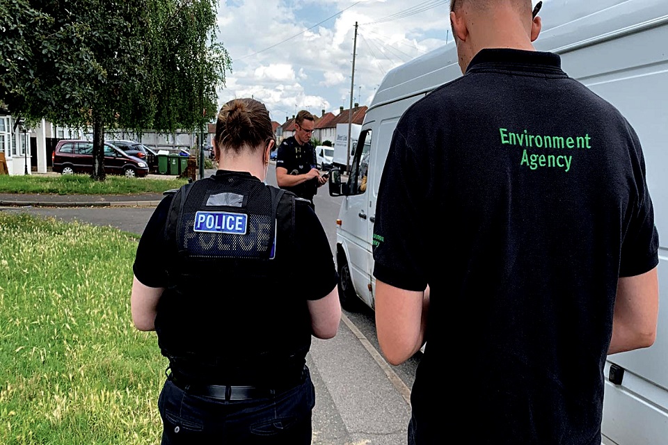 Image shows a man checking the paperwork of the driver of a white van, viewed between a police officer and an Environment Agency officer