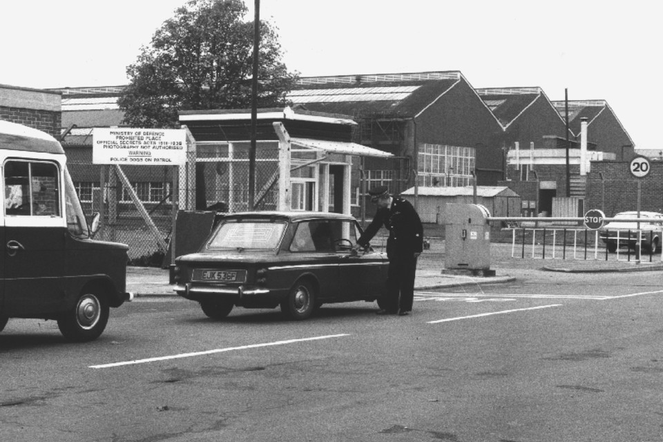 Black and white image of Ministry of Defence Police officer carrying out gate duties, alongside an arriving vehicle