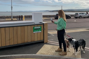 Lady walking 2 dogs holds her phone camera up to take a photo of sign on Exmouth tidal defence scheme flood gate