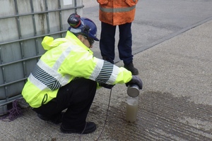 A man in a high-visibility vest is crouched down pouring dirty-looking water into a bottle.