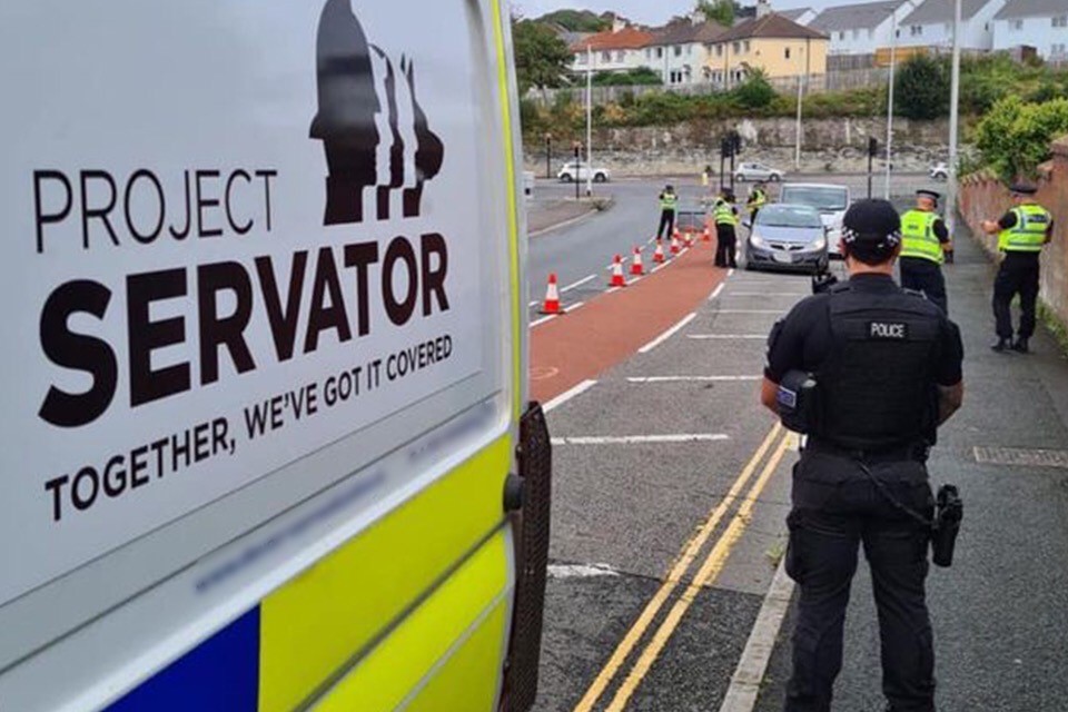 Project Servator police vehicle at roadside alongside an MDP officer, standing on the pavement, looking towards officers in high vis jackets around cars cordoned off with red traffic cones, with houses in the background and cars travelling along the road.