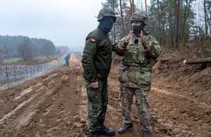 Two uniformed personnel in discussion, with a border fence to the left of them.