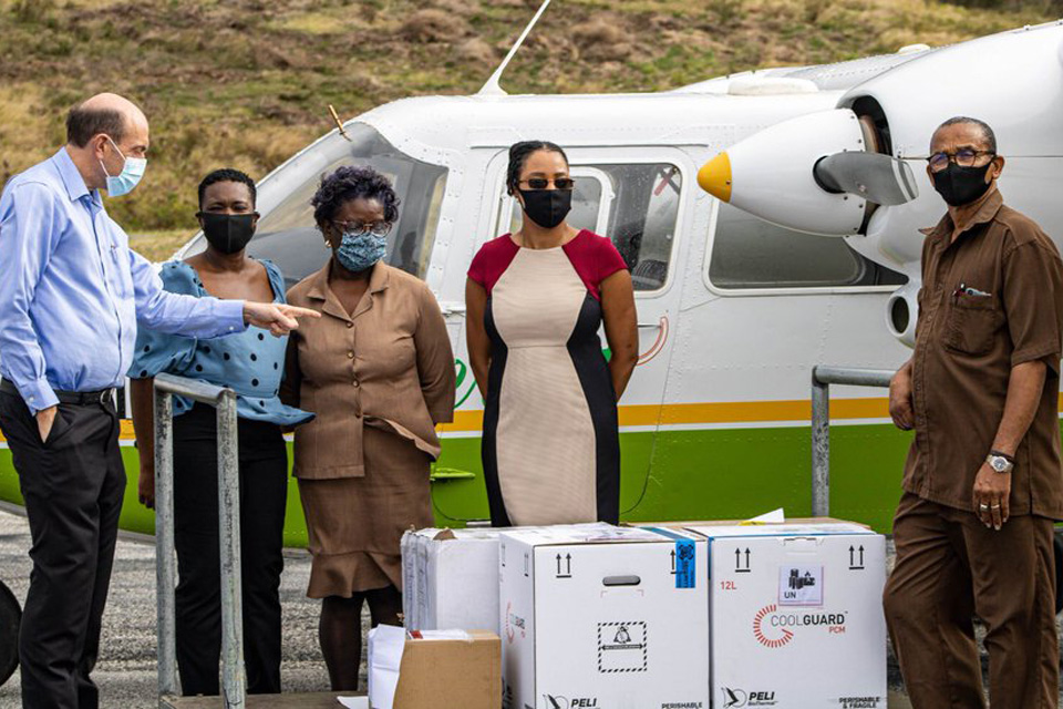 Five figures stand in front of a cargo plane, one with several boxes of Covid-19 vaccines. The figure on the left is the UK Governor of Monserrat, who is inspecting the delivery.