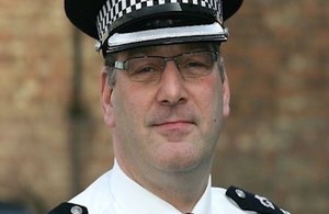 Head and shoulders of male wearing glasses and police uniform, including white shirt, black epaulettes and police hat.