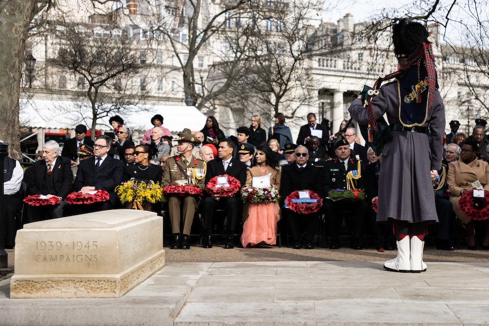 Commonwealth representatives prepare to lay wreaths at the Commonwealth Memorial Gates service in London. A piper stands in the foreground, facing them.