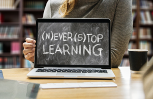 Woman sitting in library with laptop open on image of a blackboard with the words (N)ever (S)top Learning.