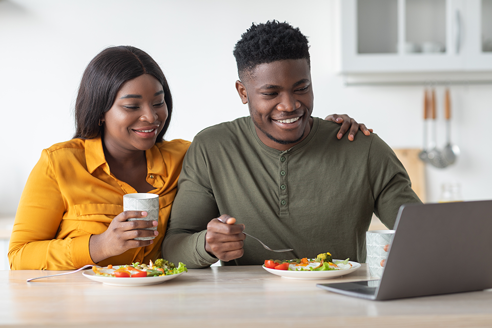 Man and woman eating breakfast in their kitchen whilst using laptop 