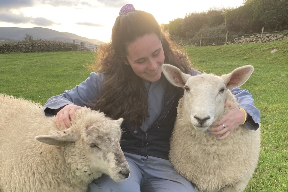 A photograph of Kayley Curtis, who is kneeling down and stroking two sheep on the farm where she lives. 