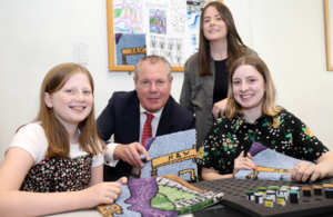 Minister Conor Burns, Jubilee design competition winner Emily McMullan, and designers Sarah Healy and Sinead Tumilty face the camera and hold up sample pieces of the designed rug.