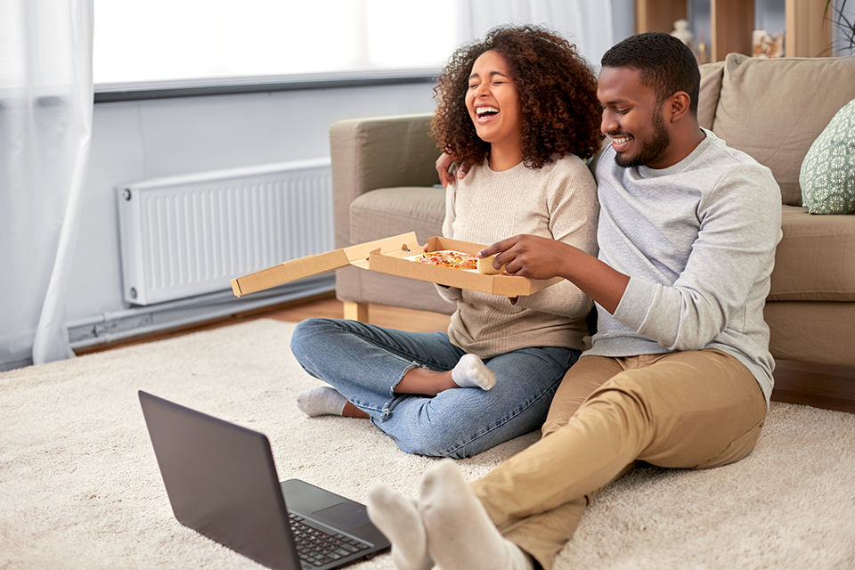 A couple watching something on their laptop, sat on the floor whilst eating a pizza
