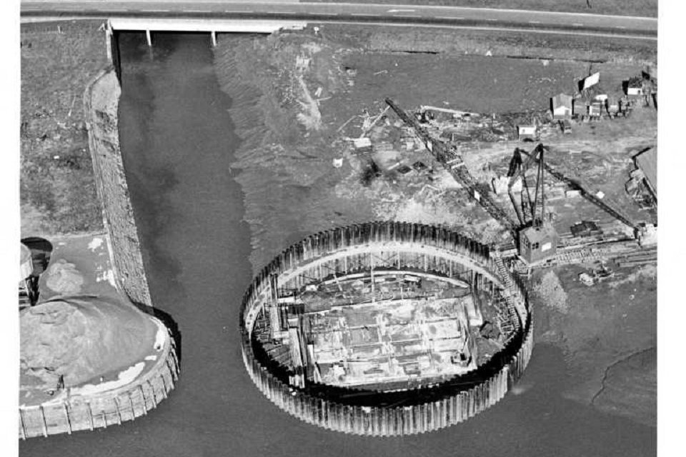 Black-and-white aerial photo of a river mouth and a large, round work site to its side 