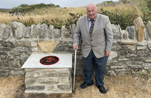 A gentleman wearing a suit stands next to a memorial plaque displaying the text: No. 4 Commando, Dieppe 1942, as well as insignia representing the unit.