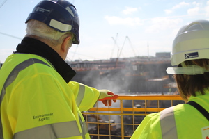 Two people in Environment Agency-branded high visibility jackets and hard hats looking at construction in the background