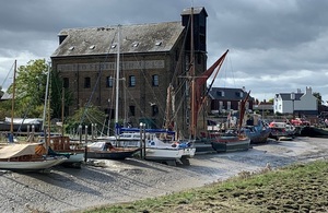 Image shows a line of sailing boats and barges resting on a river bed when the tide is out in front of an old warehouse