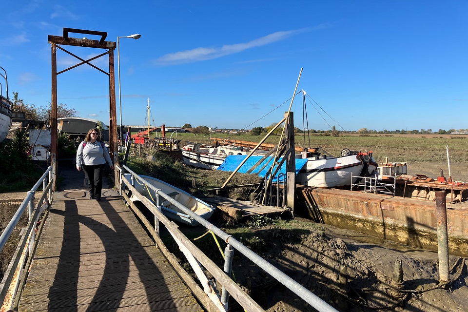 Image shows person walking towards camera over a rustic bridge to the left of the picture. Boats undergoing repair can be seen to the right