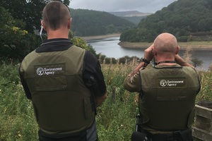 Behind two men, wearing khaki-coloured vests with Environment Agency logo, looking at a river in the distance