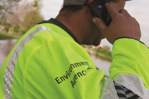 A man wearing a hard hat and a high-visibility jacket with the Environment Agency logo on his arm answers his mobile phone