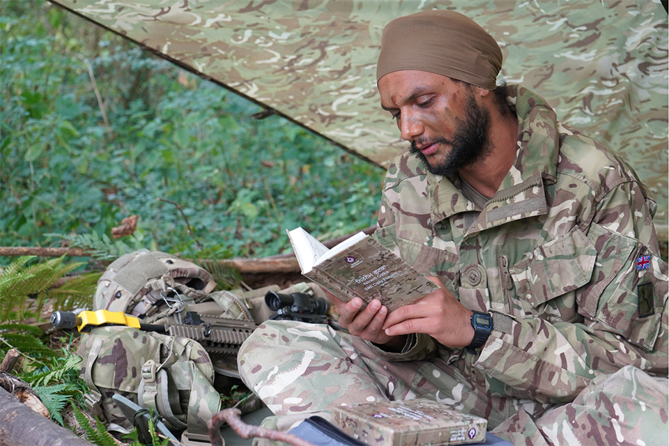 A person sitting outside on exercise in a camouflage uniform, reads the Nitnem Gutka