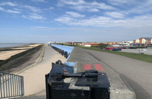 Canvey seafront with the sea, sand, parked cars and houses shown. The background is a cloudy blue sky.