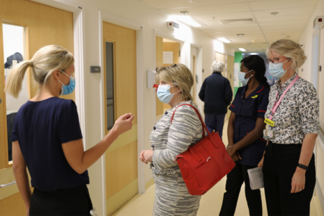 Фотография министра по делам женщин Марии Колфилд, беседующей с членами родильной бригады больницы Университетского колледжа Лондона в больничном коридоре. Все фигуры на фото носят маски.