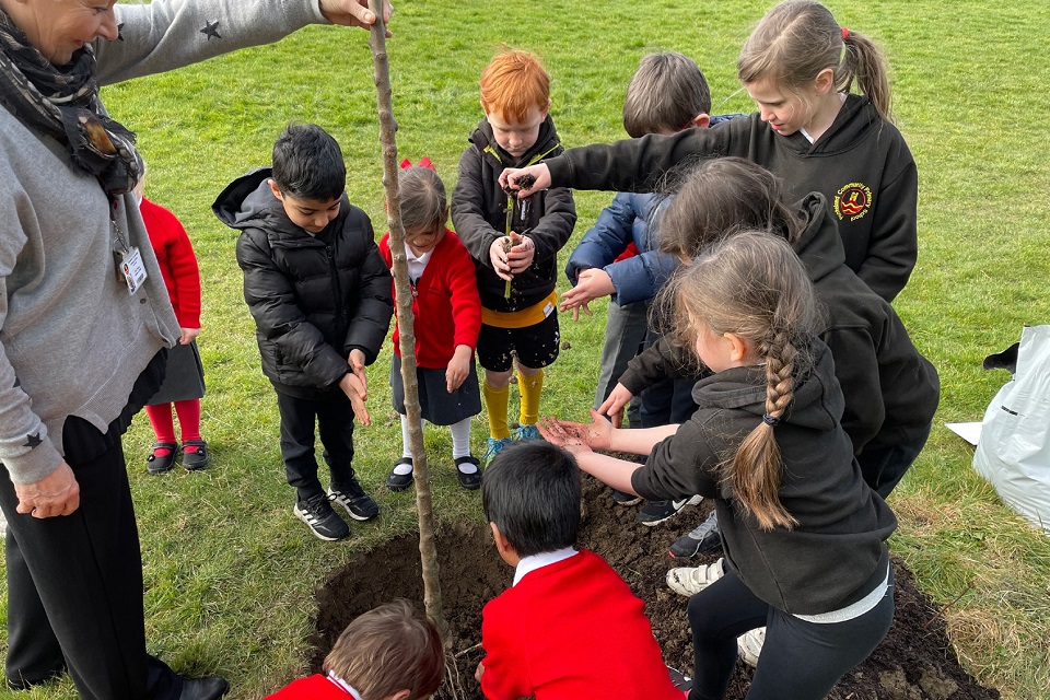 A group of primary school children, with an adult, planting a tree in a patch of ground they have dug.