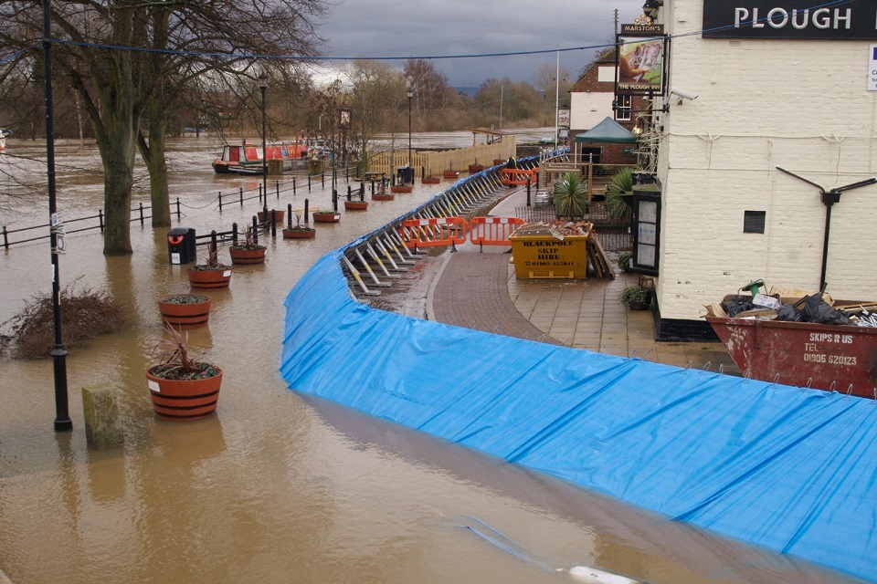 A view of a flooded urban scene. The pavements are no longer visible due to the flood waters. A temporary flood defence acts as a barrier to the flood waters reaching the buildings, including a pub called The Plough Inn.