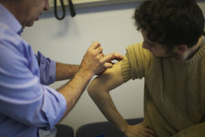 A young man has his sleeve rolled up to receive an injection in his right arm from a health professional
