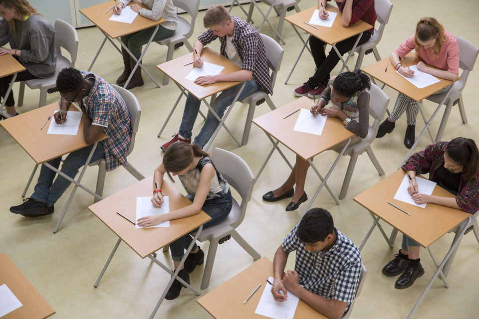 A group of students of different races and gender sitting at desks, taking an exam in a school hall