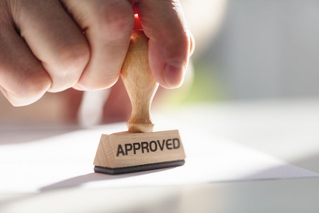 A hand marking a piece of paper with a stamp saying 'Approved'