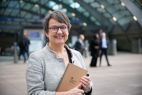 The Government Actuary, Fiona Dunsire holding a notebook with a departmental logo visible. Fiona is smiling at the viewer and is outside a London Underground station.