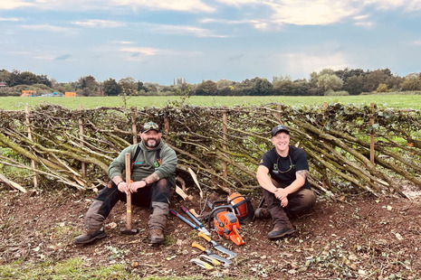 Mark and his colleague sit happily resting by a hedgerow. There's a chainsaw and other forestry tools between them.