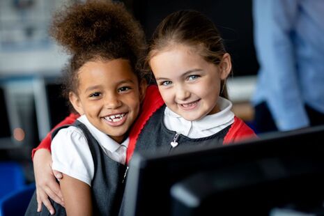A close up front-view of two best friends sitting in computer class with their arms affectionately over each other's shoulders and smiling as they look towards the camera