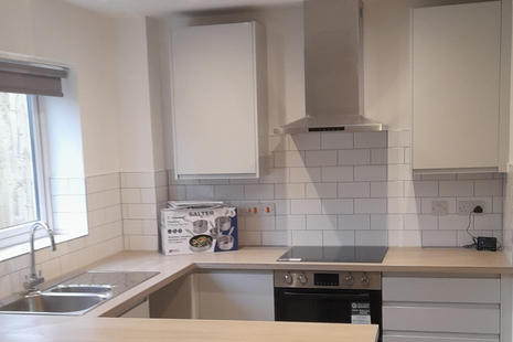 A new kitchen, with a window and sink to the left behind a countertop, and in the background, an oven, induction hob, stainless steel extractor hood, two large cupboards and a set of drawers. The kitchen is white and looks bright and airy.