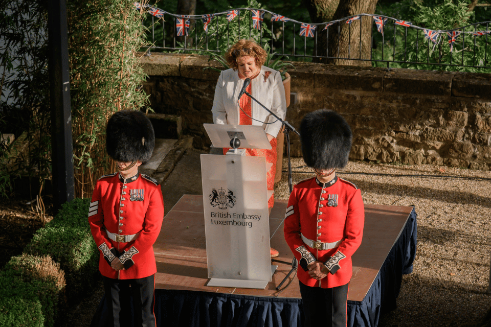 Woman in orange dress speaking on podium flanked by two guards in a red uniform and bearskins