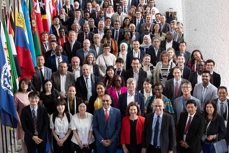 A large group of people in business attire stand together and look up at the camera. To their left is a row of flagpoles on which there are many colourful national flags.