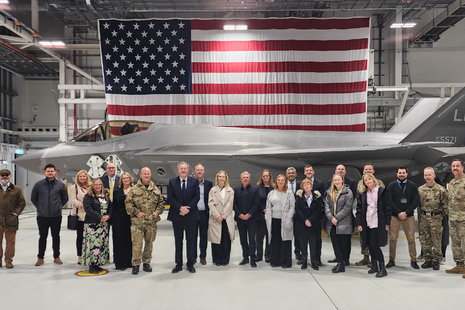 A group of people lined up in front of an F-35 aircraft inside a very large building. The people are in a mix of civilian business clothes and US Air Force uniform. A huge American flag hangs from the ceiling behind the aircraft.