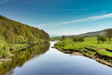 A view of the River Lune near Lancaster on a sunny day with green fields and wooded slopes
