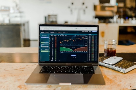 Laptop showing detailed graphs and other information in red and green. The computer is sitting on a marble table top and next to it is a notebook and phone. Credit: Unsplash.