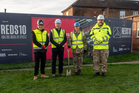 Four people stand in a line on grass smiling at the camera. Two are wearing military uniform and two are in civilian clothes, but all wear high vis, helmets and eye protection. 