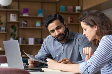 A man and woman sat at a desk in a professional setting, reviewing a document 