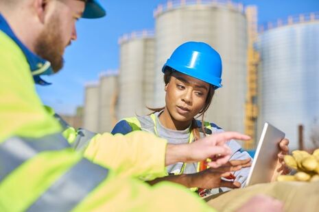 A young female student in high-vis clothing and a blue hard hat, talking to a man in similar clothing as they look at a laptop