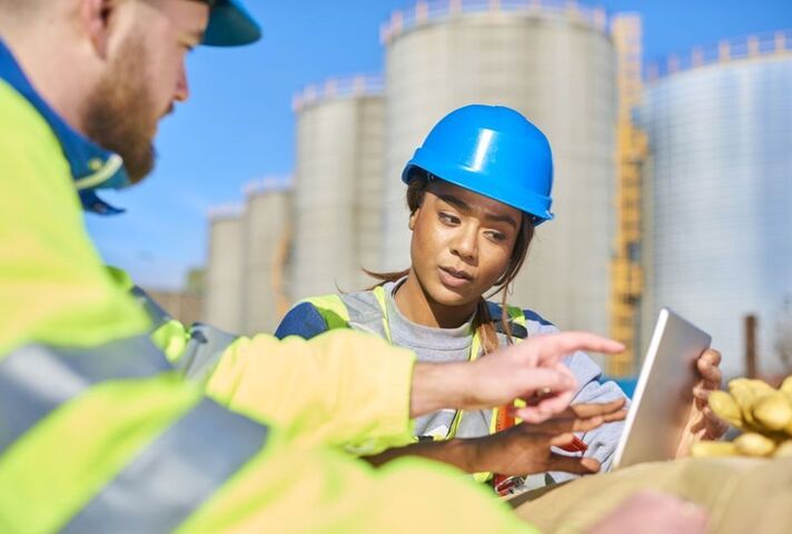 A young female student in high-vis clothing and a blue hard hat, talking to a man in similar clothing as they look at a laptop