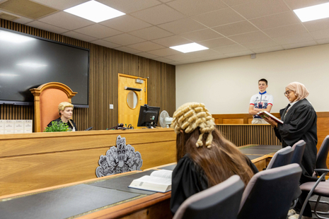 A courtroom scene with young people acting as a judge, two barristers, and a witness standing within a wooden witness box. Various books and a monitor are visible.